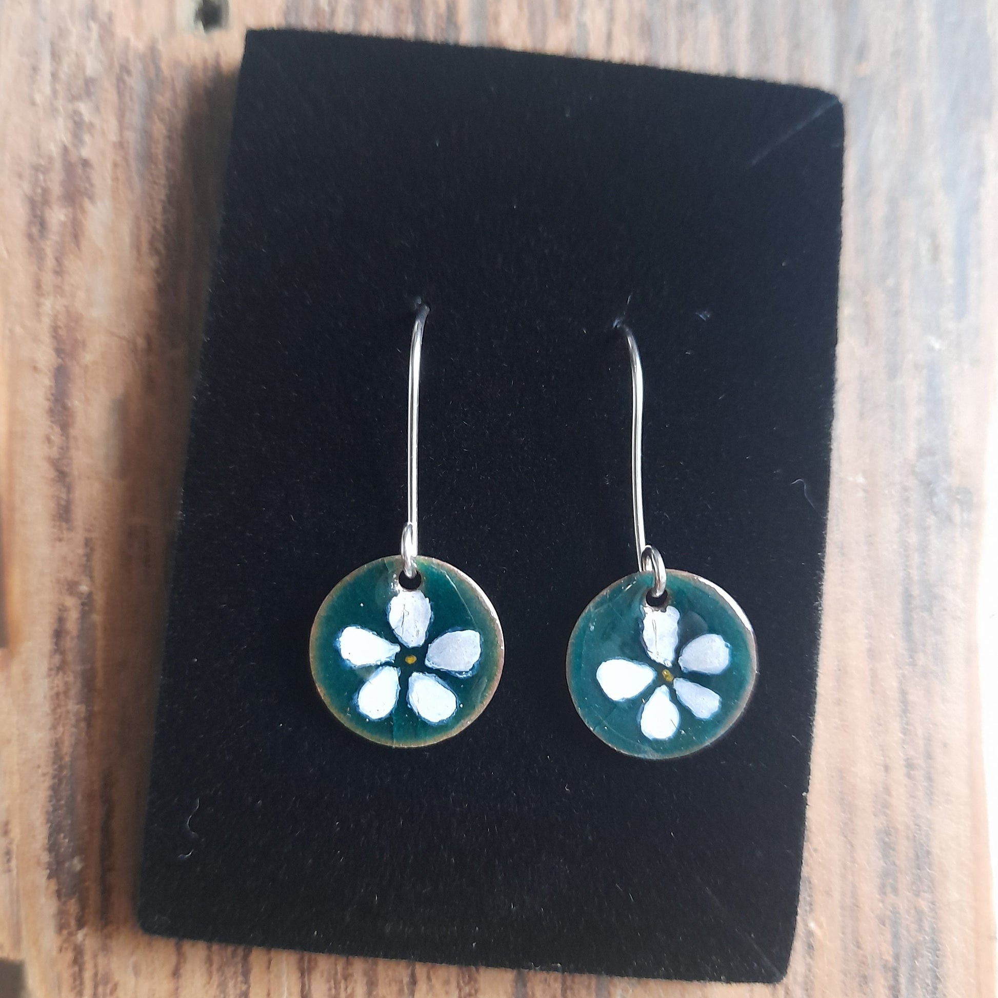 Circular disc earrings in Teal, with a white handpainted flower on silver earwires. On black background 
