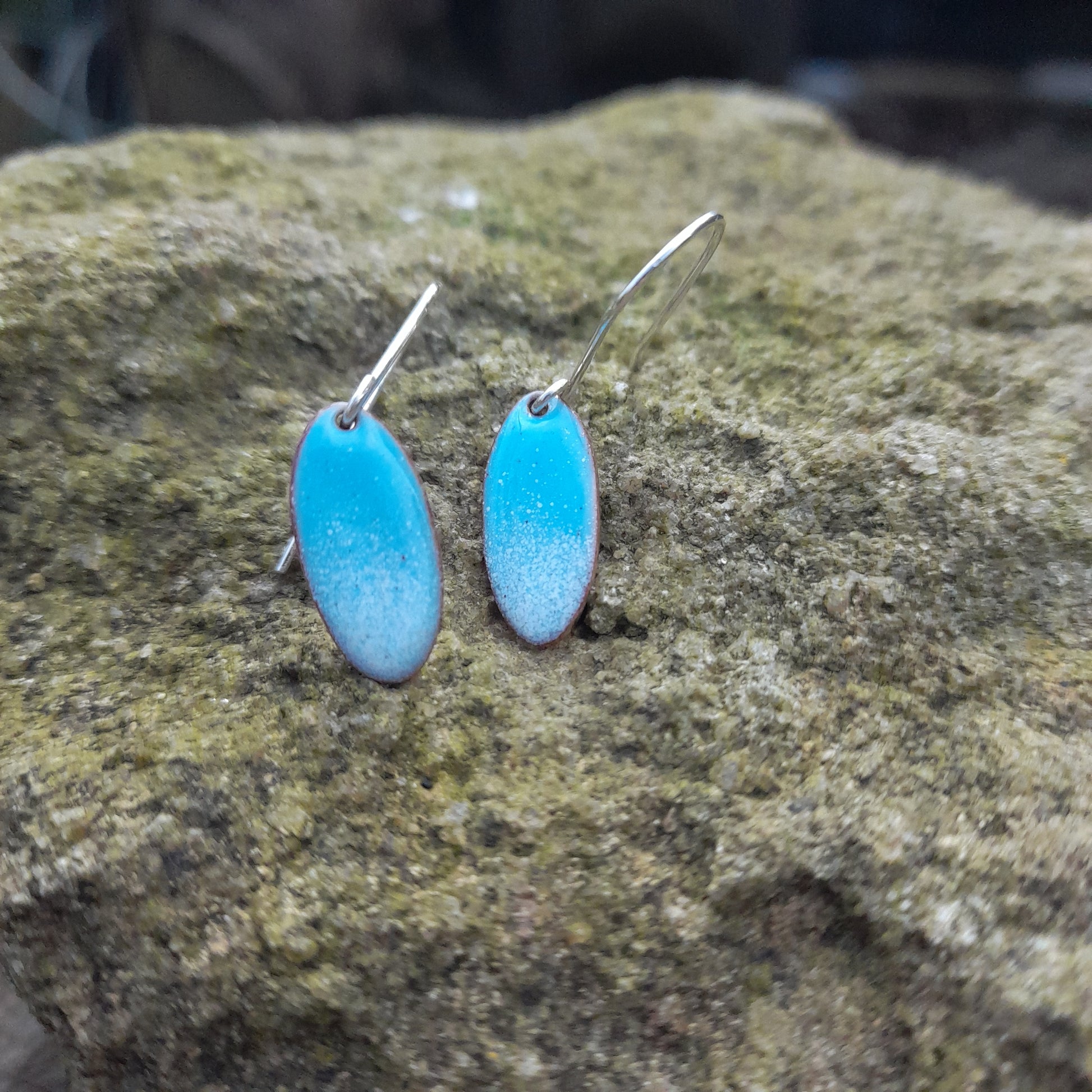 Pale blue oval copper and enamel earrings with silver handmade earwires. Slight graduation to pale blue at the bottom, displayed on a stone outside. 