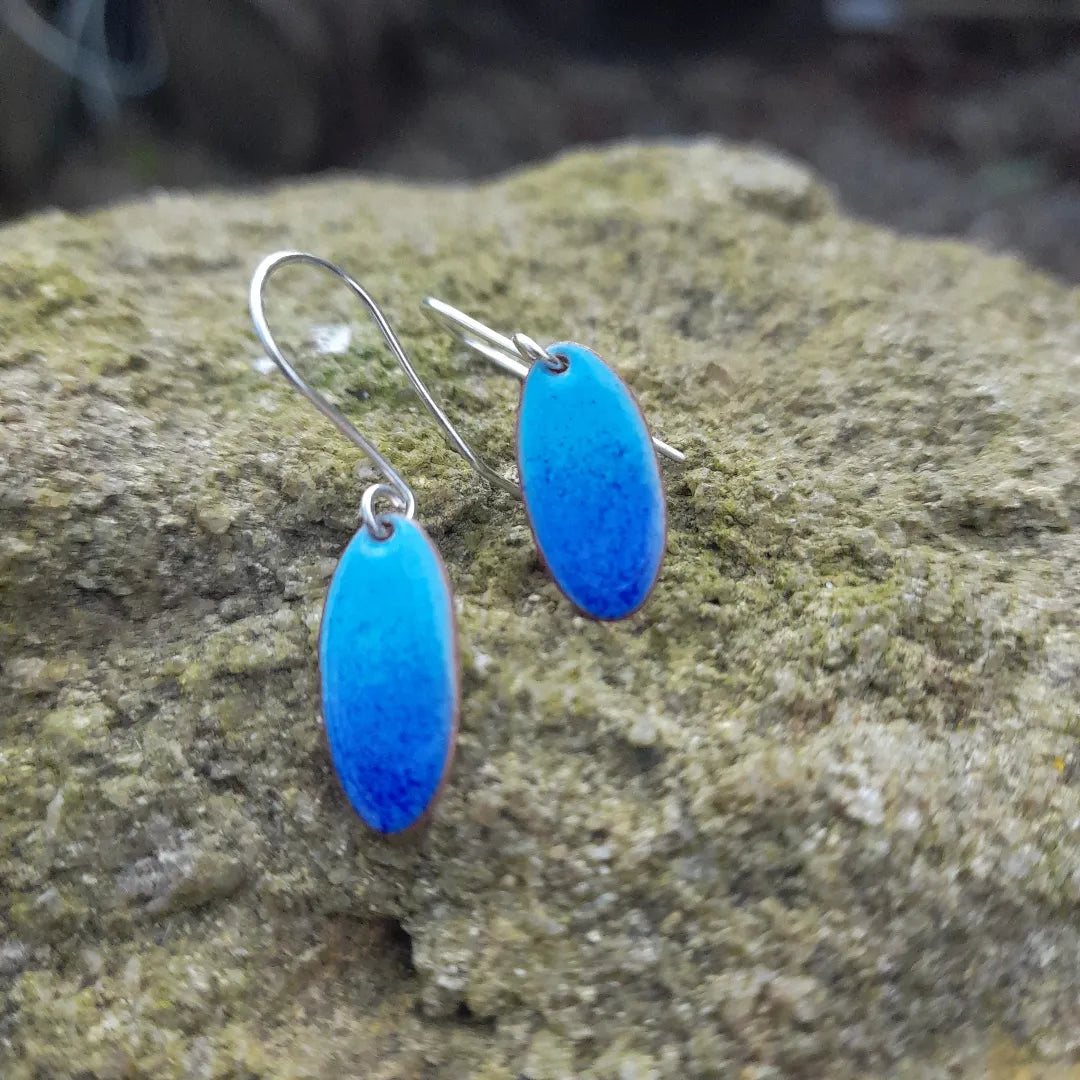 Pale blue oval copper and enamel earrings with silver handmade earwires. Slight graduation from pale blue at the top to a darker blue at the bottom, displayed on a stone outside. 