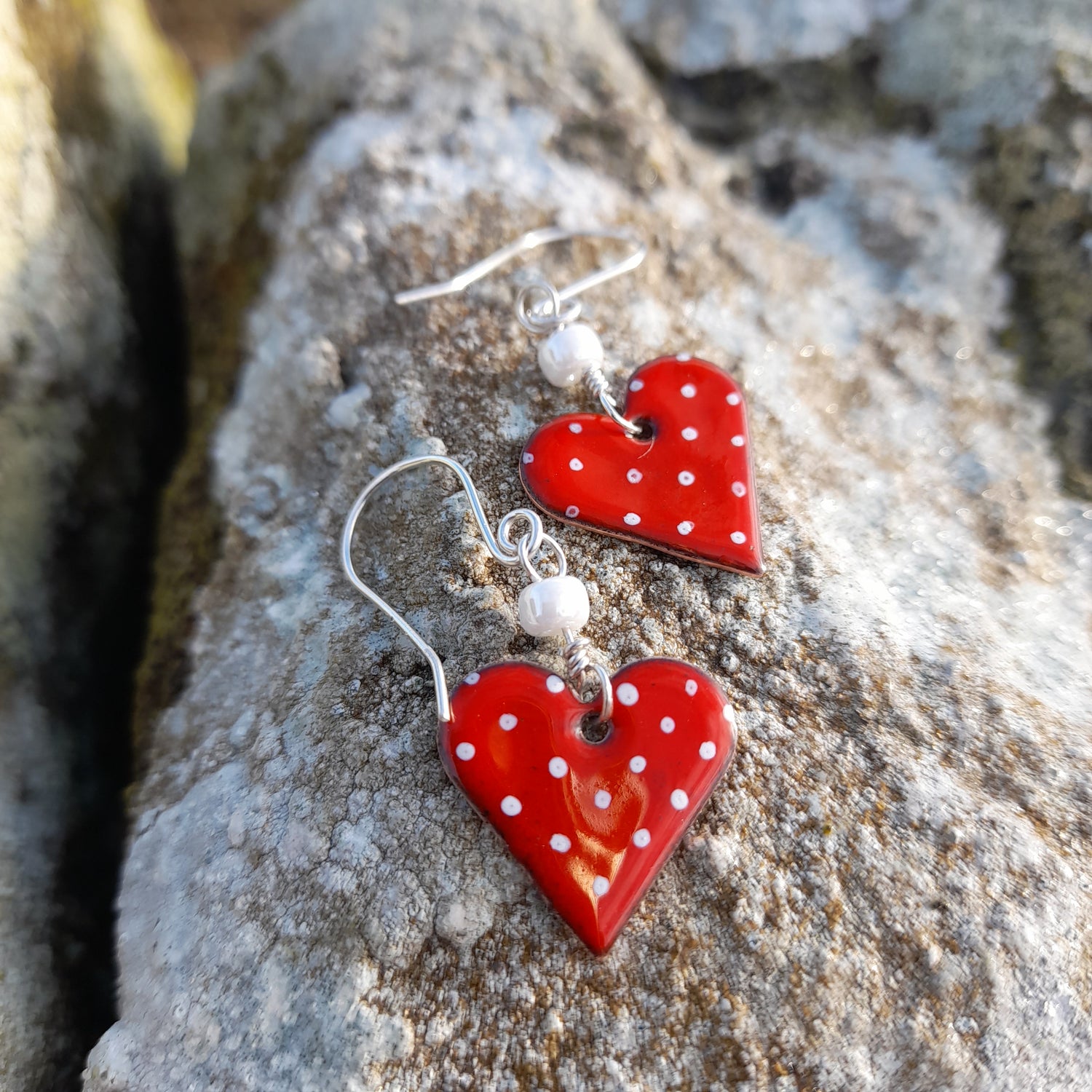 red polka dot handmade heart earrings, on silver wire with white bead. They are photographed on a white rock in daylight. 