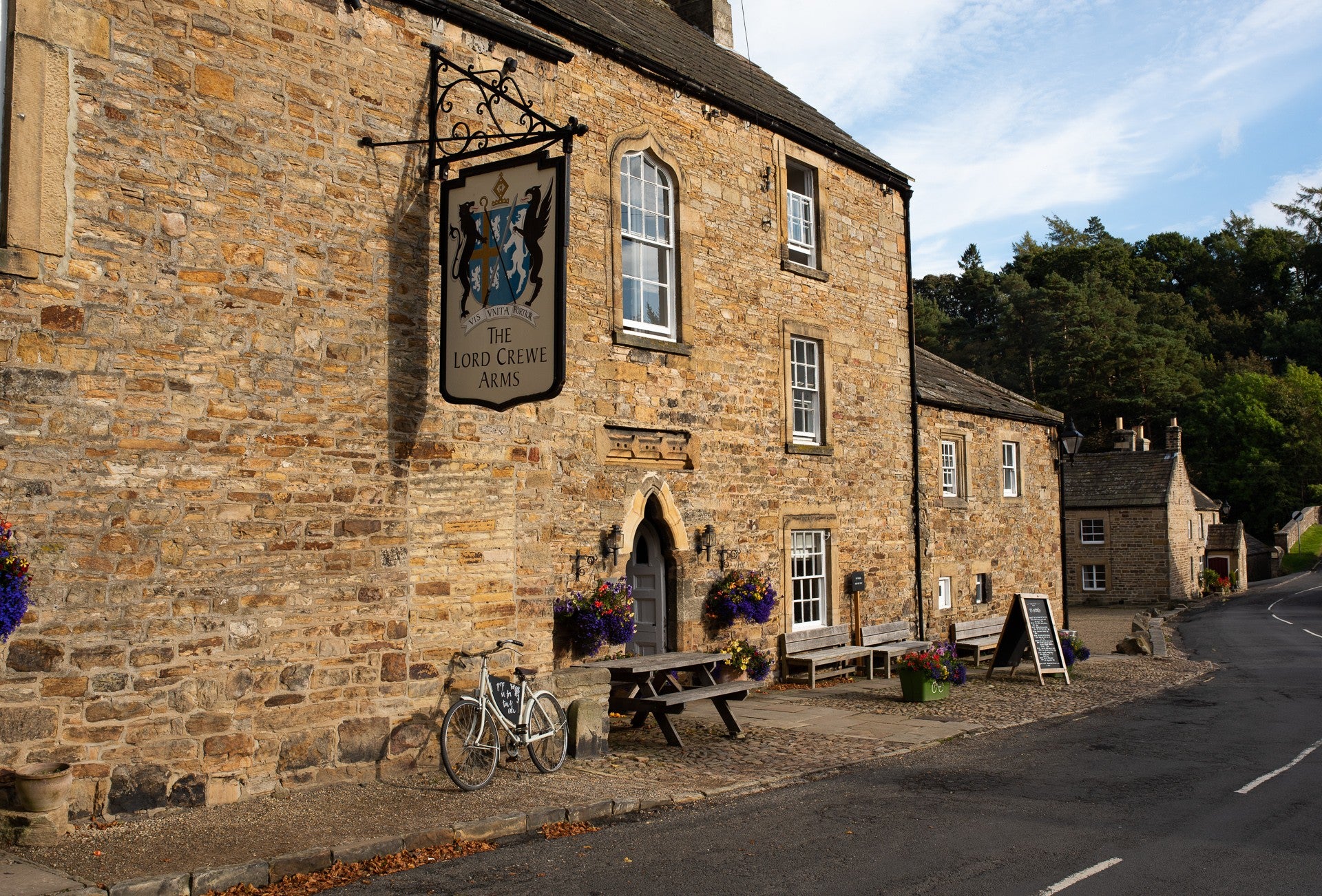 The picture shows the front profile of the Lord Crewe Arms Hotel in Blanchland on a sunny day. There are trees in the background and pub benches at the front.